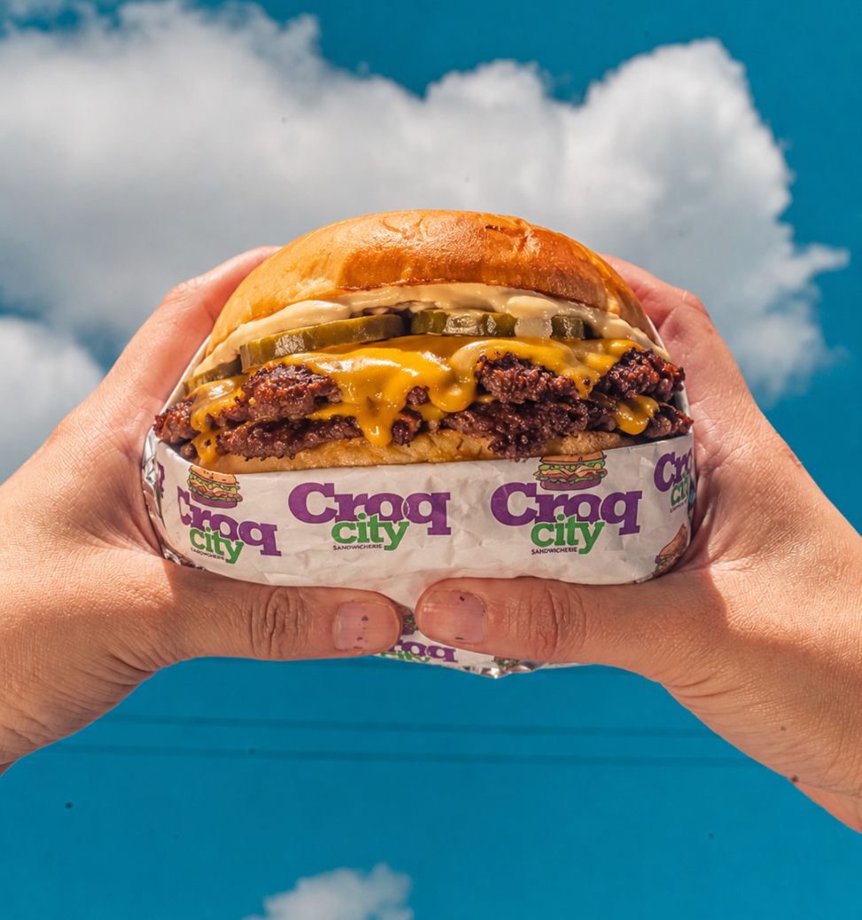 Smash burger CroqCity tenu à deux mains sur fond de ciel bleu